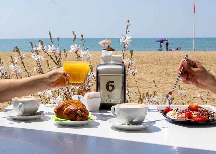 Colazione in spiaggia: caffè, succo, cornetto, mare sereno, atmosfera rilassata.