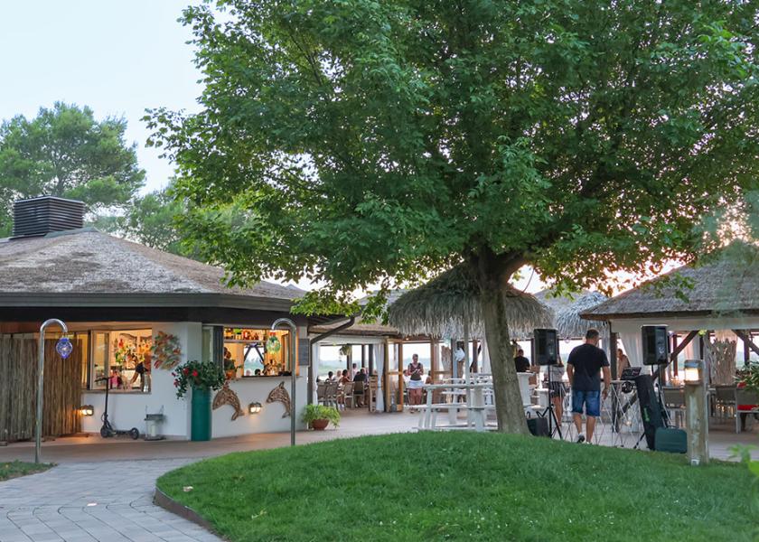 Ristorante all'aperto con tetto in paglia, circondato da alberi e persone sedute.